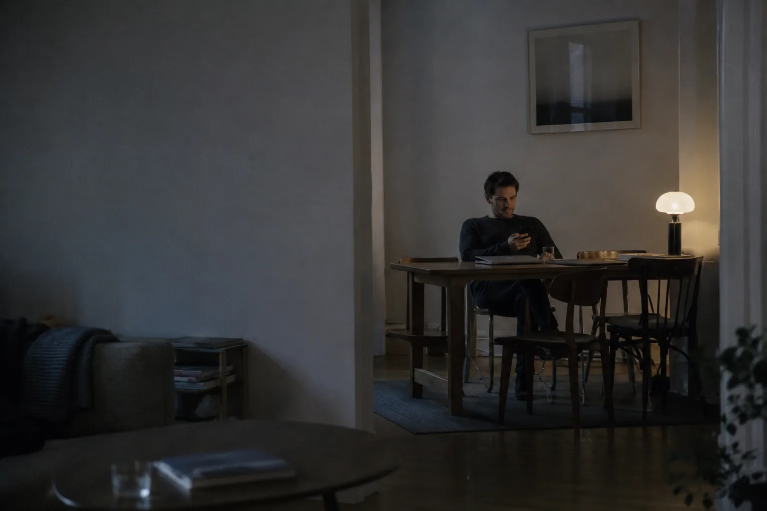 A person sitting alone at a table in a dim living room in the evening, reading something on a phone, seen from across the room.