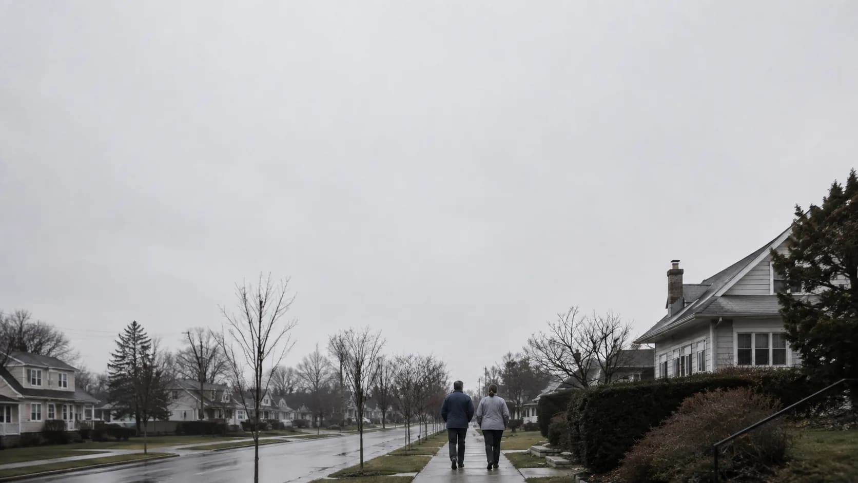 Two people in their fifties walking away from camera along a suburban sidewalk on an overcast morning, under a wide flat gray sky.